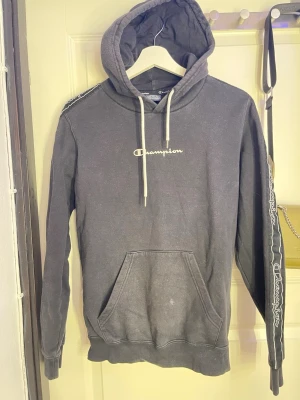 Svart Champion hoodie med logga - Säljer en svart hoodie från Champion med vit broderad logga på bröstet och vita snören i huvan. Ärmarna har band med Champion-tryck längs sidorna och en stor magficka framtill. Hoodien är i mjukt bomullsmaterial och har ribbade muddar vid ärmslut och nederkant.