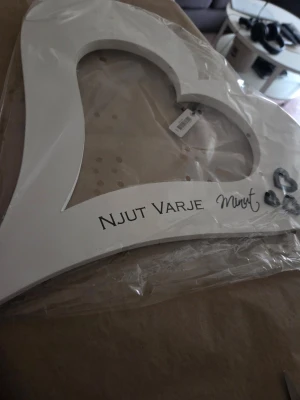 Vitt hjärtformat väggord i trä - Dekorativt väggord i vitt trä med hjärtform och texten 'NJUT VARJE minut'. Perfekt som inredningsdetalj i hemmet. Produkten har en liten metallfjäril som extra detalj och mäter cirka 40 cm på längsta sidan.