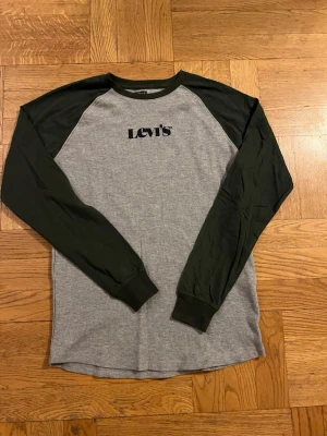 Grå långärmad topp från Levi's - Grå långärmad topp från Levi's med svarta ärmar och svart logga på bröstet. Klassisk raglanmodell med rund halsringning. Tillverkad i mjuk bomull, perfekt för en avslappnad stil.