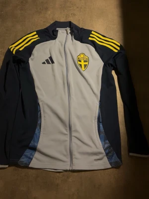 Svensk landslagsträningströja Adidas - Träningströja från Adidas i Sveriges landslagsfärger. Grå front med mörkblå ärmar och axlar, gula detaljer och tre ränder på axlarna. Broderad Sverige-logga på bröstet och Adidas-logga. Hel dragkedja och hög krage. Perfekt för fotbollsträning.