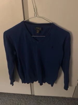 Blå V-ringad tröja Polo Ralph Lauren - Snygg blå V-ringad tröja från Polo Ralph Lauren i mjuk pima bomull. Klassisk design med diskret logga på bröstet och ribbade muddar vid ärmslut och nederkant. Perfekt för en stilren och enkel look.