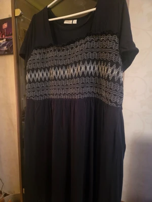 Svart klänning med mönster från Zizzi - Svart långklänning från Zizzi med korta ärmar och vitt geometriskt mönster över bröstet. Klänningen har rund halsringning och är tillverkad i mjukt bomullstyg. Perfekt för dig som gillar en avslappnad och bekväm stil.