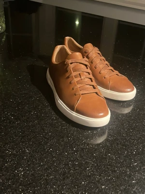 Bruna Clarks sneakers i skinn - Snygga bruna sneakers från Clarks Unstructured. Modellen heter UN COSTA LACE. Skon är gjord  i Läder med vita sulor och matchande bruna snören. Skon har en klassisk låg modell med rund tå och stilren design som passar till det mesta. Perfekta för dig som gillar en stilren och bekväm sko inför sommaren.