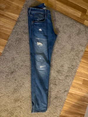 Replay anbass jeans  - Säljer ett par blå s jeans med slitningar på framsidan. Jeansen har klassisk femficksdesign och normalhög midja. Perfekta för dig som gillar en avslappnad och trendig stil. Storlek W31/L32