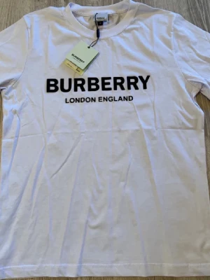 Vit Burberry t-shirt med Burberry logga - Snygg vit t-shirt från Burberry med klassisk svart logga och texten 'London England' på bröstet. T-shirten har rund hals och korta ärmar, tillverkad i mjuk bomull för en skön känsla. Perfekt för dig som gillar stilrena och ikoniska plagg. Priset kan diskuteras !