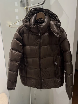 Brun pufferjacka från Massimo Dutti - Snygg brun pufferjacka från Massimo Dutti med hög krage och avtagbar huva. Jackan har två fickor med dragkedja och quiltad design. Perfekt för kalla dagar och ger en modern, puffig siluett. Stängs med dragkedja framtill.