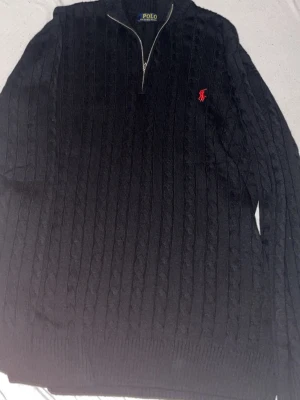 Svart kabelstickad tröja Polo Ralph Lauren - Kabelstickad svart tröja från Polo Ralph Lauren med halv dragkedja och röd broderad logga på bröstet. Tröjan har ribbade muddar och hög krage, perfekt för lager på lager. Klassisk och stilren design som funkar till det mesta.