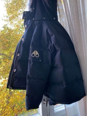  Moose Knuckles - Snygg svart dunjacka från Moose Knuckles med silverfärgad logga på ärmen och stor huva med fluffig pälskant. Jackan har dragkedja, knappar och flera fickor. Perfekt för kalla dagar och riktigt varm.