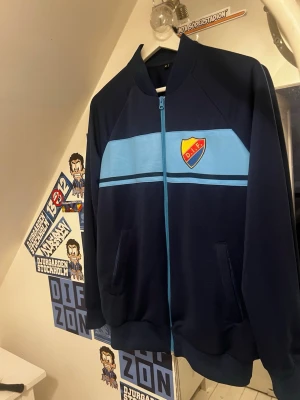 Blå DIF ziptröja med klubbmärke - Mörkblå ziptröja från Djurgårdens IF med ljusblåa detaljer och klubbmärke på bröstet. Tröjan har hel dragkedja framtill, två fickor och ribbade muddar. Perfekt för dig som vill visa vilket lag du håller på.