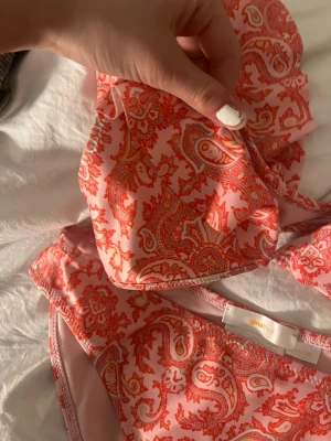 Rosa paisleymönstrad bikini från Småfolk - Säljer en snygg bikini från Gina tricot i rosa med orange paisleymönster. Bikinin har klassisk trekantsmodell på toppen och matchande trosor. Materialet är mjukt och stretchigt, perfekt för sommaren och strandhäng.