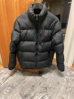 Svart dunjacka från Ralph Lauren - Snygg svart puffer dunjacka från Ralph Lauren med hög krage och dragkedja framtill. Jackan har det klassiska röda Polo-logot på bröstet och två sidofickor med dragkedja. Perfekt för kalla dagar och ger en clean, modern look.Liten defekt, kolla sista bild. 