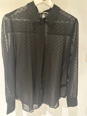 Svart transparent skjorta med hjärtan storlek 38 - Säljer en svart transparent skjorta med små hjärtmönster. Skjortan har klassisk krage, knappar framtill och långa ärmar. Perfekt för dig som vill ha en unik och trendig look med lite extra detaljer.