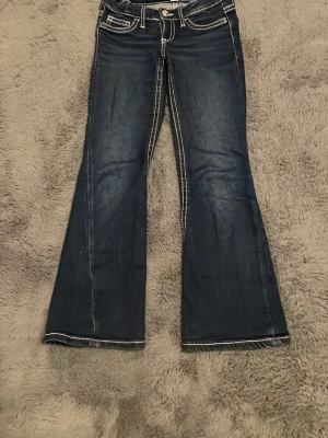 Mörkblå bootcut jeans  - Säljer ett par mörkblå bootcut jeans med vita kontrastsömmar och snygga detaljer på bakfickorna. Jeansen har låg midja och klassisk femficksdesign. Perfekta för dig som gillar en retroinspirerad look och vill ha ett par jeans som sticker ut.
