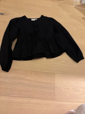 Svart blus med knyt från Gina Tricot - Supersnygg svart blus från Gina Tricot med långa ärmar och volang nedtill. Blusen har knytning framtill och en lös, flowy passform som ger en chill vibe. Perfekt att styla med jeans eller kjol för en trendig look. Men har ett hål på vänstra axlen men går enkelt att laga. Har aldrig använt den.