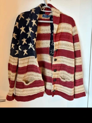 Stickad cardigan Polo Ralph Lauren XS - Säljer en unik stickad kofta från Polo Ralph Lauren i storlek XS. Koftan har amerikanskt flaggmönster med röda, beige och blå ränder samt stjärnor. Den har knappar framtill, två fickor och är gjord i grovstickat material. Perfekt för dig som vill sticka ut!