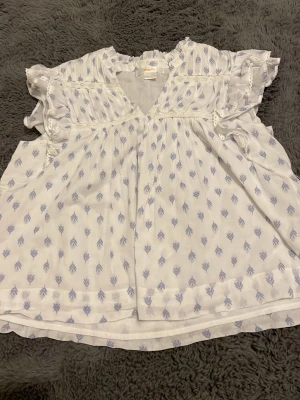 Vit blus med volanger och blått mönster - Söt vit blus med ljusblått bladmönster och volangdetaljer vid ärmar och axlar. Blusen har v-ringning, spetsinslag och är tillverkad i ett tunt, luftigt tyg som känns som bomull. Perfekt för en avslappnad och trendig look.