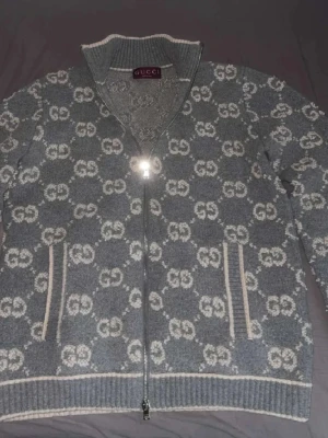 Grå stickad ziptröja  - Grå stickad tröja med dragkedja från Gucci. Tröjan har ett helprintat GG-mönster i vitt, ståkrage och två fickor framtill med vita detaljer. Ribbstickade muddar och nederkant ger en snygg finish. Perfekt för dig som gillar lyxiga streetwear vibes.