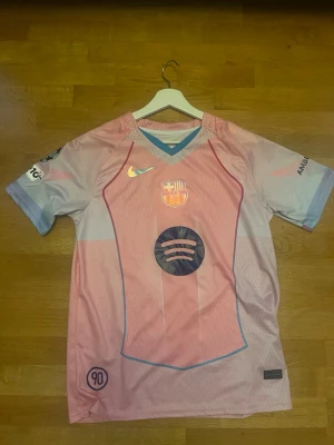 FC Barcelona rosa matchtröja Nike - Säljer en rosa och ljusblå FC Barcelona matchtröja från Nike med klubbmärke, Spotify-logga och UNHCR-tryck. Tröjan har korta ärmar, V-ringning och detaljer i blått. Perfekt för fotbollsfans som vill sticka ut på planen.