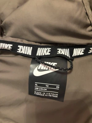 Nike pufferjacka med huva barn XL - Snygg pufferjacka från Nike i färgerna svart, beige och brun. Jackan har huva, dragkedja framtill och två sidofickor. Perfekt för kalla dagar och har en sportig look med klassisk Nike-logga på bröstet.