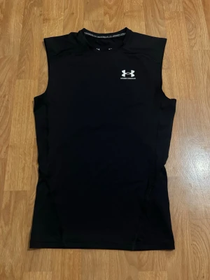 Svart kompressionstopp Under Armour - Svart ärmlös kompressionstopp från Under Armour med HeatGear-teknologi. Toppen har en rund halsringning och en vit logga på bröstet. Perfekt för intensiva träningspass och sportaktiviteter.