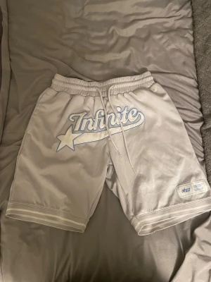 Vita Infinite shorts med snörning - Snygga vita shorts från Infinite med ljusblå logga och stjärndetalj framtill. Shortsen har elastisk midja med snörning, sidofickor och en bakficka. Perfekta för en avslappnad stil.