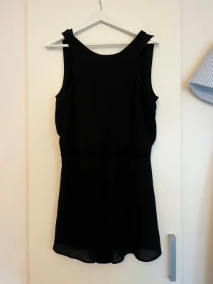 Svart ärmlös jumpsuit med knytband - Svart jumpsuit från Gina Tricot med breda axelband och knytband i ryggen. Modellen är ärmlös och har en rund halsringning. Det är ett tunt och lätt tyg som ger ett snyggt fall. I storlek 38.