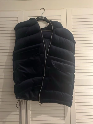 Mörkblå dunväst med huva - Snygg mörkblå dunväst med stor huva och dragkedja framtill. Västen har två stora fickor på framsidan och en quiltad design som ger en cool och modern look. Perfekt för lager-på-lager-stil under kyliga dagar.