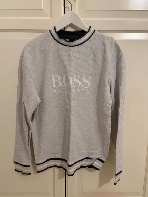 Grå sweatshirt från Hugo Boss - Snygg grå sweatshirt från Hugo Boss med stor logga framtill. Tröjan har svarta och vita ränder vid krage, ärmslut och nederkant. Perfekt för en avslappnad och trendig stil.