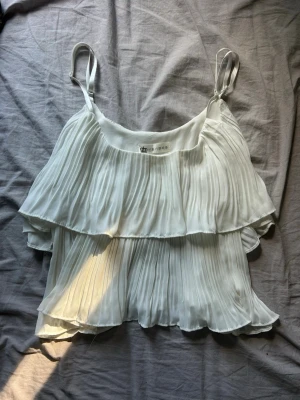 Vit plisserad linnetopp med volanger - Säljer en superfin vit linnetopp med tunna axelband och dubbla lager volanger. Toppen är plisserad för en luftig och flowy känsla, perfekt för varma dagar. Passar dig som gillar en romantisk och trendig stil.