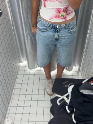 Ljusblå denim bermudashorts - Säljer ett par ljusblå bermudashorts i denim med klassisk femficksdesign och knappgylf. Shortsen har en relaxed, loose passform och råa kanter vid bensluten. Perfekta för en chill och avslappnad stil.