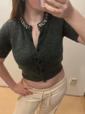 Mörkgrå kofta med fina detaljer - Kort, mörkgrå kofta med korta ärmar och knappar framtill. Kragen är dekorerad med pärlor och glittriga stenar som ger en elegant touch. Materialet ser mjukt och fluffigt ut, perfekt för att lyfta en outfit.