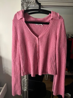 Rosa ribbad kofta med knappar - Säljer en söt rosa ribbad kofta med v-ringning och krage. Koftan har långa ärmar, fyra matchande knappar framtill och är croppad i modellen. Perfekt att styla med jeans eller kjol för en trendig look.