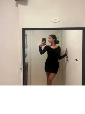 Svart tight långärmad klänning - Säljer en svart, figurnära klänning med lång ärm och djup urringning både fram och bak. Klänningen är kort och har en enkel, stilren design som passar perfekt för en trendig look. Materialet är stretchigt och följsamt för en snygg passform.