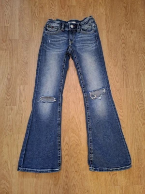 Blå bootcut jeans med slitningar - Snygga blå bootcut jeans med slitningar på båda knäna och broderade detaljer på sopckfickorna bak. Jeansen har klassisk femficksmodell och en lätt utsvängd passform nedtill. Perfekta för en avslappnad och trendig look. Storlek 158 motsvarande 34.