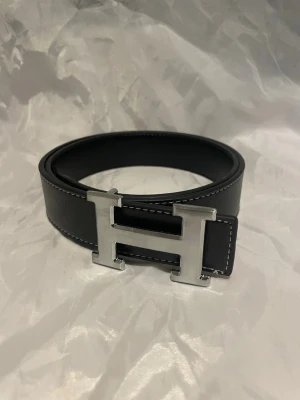 Svart bälte med H-spänne - Snyggt svart bälte i läder med ett stort, silverfärgat H-spänne framtill. Bältet har vita kontrastsömmar längs kanterna. 110cm. Pris går att diskutera 