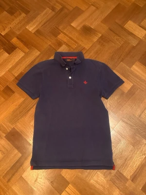 En mörkblå piké från Morris  - Klassisk marinblå pikétröja med krage och två knappar framtill. Tröjan har en liten röd broderad logga på bröstet och röda detaljer på insidan av kragen och slitsarna. Perfekt till jeans eller shorts för en clean look. Skulle säga att den passar runt 163cm. Skriv vid funderingar! 