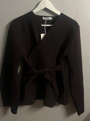 Svart omlottopp med knytband - Stilren svart omlottopp från J'aime med långa ärmar och knytband i midjan. Toppen har en v-ringad omlottdesign som ger en snygg siluett och passar perfekt till både jeans och kjol. Materialet känns mjukt och bekvämt.