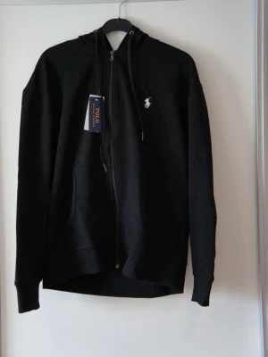Svart zip hoodie från Polo Ralph Lauren - Snygg svart hoodie med dragkedja från Polo Ralph Lauren. Klassisk design med vit broderad logga på bröstet, huva med dragsko och kängurufickor framtill. Tillverkad i mjukt bomullsmaterial som känns skönt mot huden. Perfekt för en avslappnad och stilren look. ❗️OBS❗️ - Skriv till mig innan köp! 🙌🏼