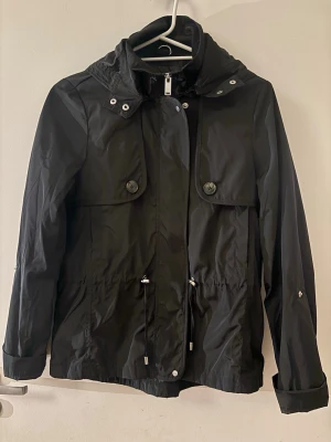 Svart vindjacka från Vero Moda - Snygg svart vindjacka från Vero Moda med huva, stora fickor med knappar och dragsko i midjan. Jackan har dragkedja framtill och långa ärmar.  Enkel att matcha med olika outfits.