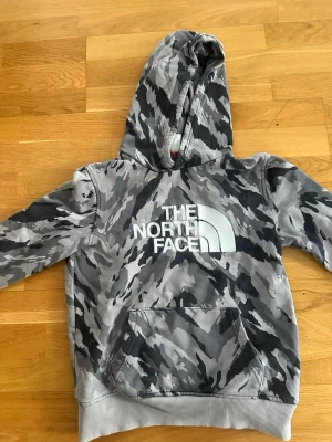 Camouflage hoodie från The North Face - Snygg hoodie från The North Face i grått och svart camouflagemönster. Stor logga framtill, huva och känguruficka. Tillverkad i mjukt material med ribbade muddar vid ärmslut och nederkant. Perfekt för en avslappnad streetwear-look. Passar dig som är mellan 145/160
