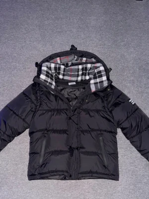 Svart pufferjacka från Burbérry - Snygg svart pufferjacka från Burbérry med klassiskt rutigt foder i huvan. Jackan har dragkedja framtill, två snedställda fickor och en patch med Burbérry London England på ärmen. Perfekt för kalla dagar och riktigt trendig look.