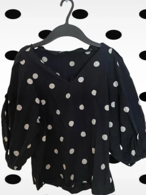 Svart prickig blus med v-ringning - Trendig svart blus med vita polkaprickar och v-ringad hals. Blusen har en lätt puff. Tillverkad i ett mjukt, tunt tyg som ger en avslappnad och luftig känsla. Originalpris: 300 kr