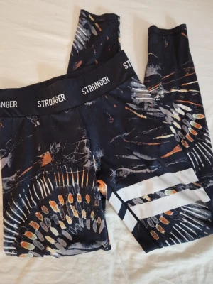 Mönstrade tights från Stronger - Snygga svarta tights från Stronger med färgstarkt abstrakt mönster i orange, vitt och gult. Bred resår i midjan med logga runtom och vita ränder på ena benet. Perfekta för träning eller gymmet. Byxorna är i fint använt skick. Hund finns I hemmet. Märkt med E.B på lappen på insidan.