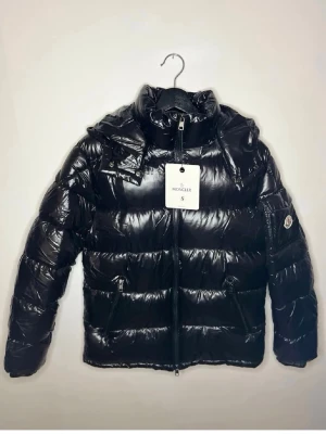 Svart puffer dunjacka från Moncler - Snygg svart dunjacka från Moncler med glansig finish och klassisk pufferdesign. Jackan har huva, dragkedja framtill, två sidofickor med dragkedja och en ärmficka med Moncler-logga. Perfekt för kalla dagar och riktigt trendig look. NFC finns på armen. Monclear påsen ingår också. Vid frågor eller fler bilder skriv gärna, priset går att diskutera