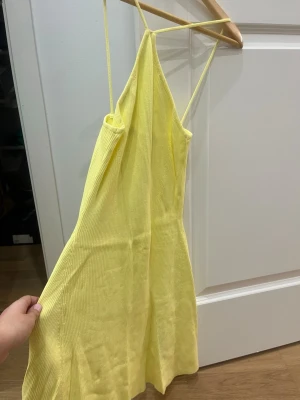 Gul ribbad kortklänning med smala band - Säljer en söt gul kortklänning med smala axelband och ribbad struktur. Klänningen har en lätt utsvängd passform och är perfekt för varma dagar. Materialet känns mjukt och stretchigt, vilket gör den bekväm att bära.