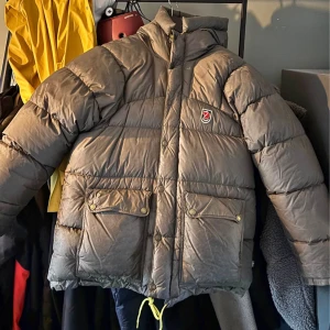 Brun dunjacka från Fjällräven - Fjällräven, grön i äldre modell från 2008/2009. Använd i någon säsong i princip och har sedan dess hängt i förrådet. Kan behöva kemtvätt. Har en reva baktill längst ner till vänster om jackan(4,5x4,5 cm) dock endast yttertyget och inte i dunet! Passar både män och kvinnor.