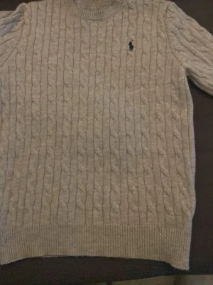 Grå kabelstickad tröja från Polo Ralph Lauren - Kabelstickad grå tröja från Polo Ralph Lauren med rund halsringning och svart broderad logotyp på bröstet. Tröjan har ribbade muddar vid ärmslut och nederkant, perfekt för en klassisk och stilren look.