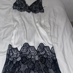 Vit satinklänning med svart spets - Supersnygg vit satinklänning med svarta spetsdetaljer upptill och nertill. Klänningen är ärmlös och har en figurnära passform med v-ringad spets vid bysten. Perfekt för dig som gillar en elegant och chic look. storleken är 34 men jag upplever att den känns mycket större än en 34.