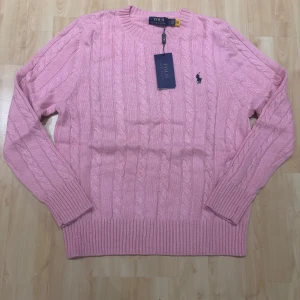 Rosa kabelstickad tröja Polo Ralph Lauren - Supersnygg rosa kabelstickad tröja från Polo Ralph Lauren med klassisk rund halsringning och broderad logga på bröstet. Tröjan har långa ärmar och ribbade muddar för en snygg passform. Perfekt för lager på lager under kyliga dagar.