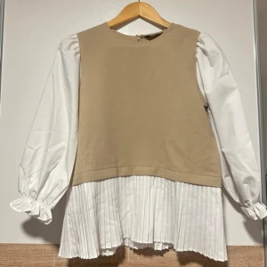 Beige och vit blus med plisserad nederdel - Snygg blus i beige och vitt med puffiga långärmar och plisserad nederdel. Toppen har rund halsringning och en cool kontrast mellan den släta beigea överdelen och de vita detaljerna. Perfekt för dig som gillar trendiga och unika plagg.